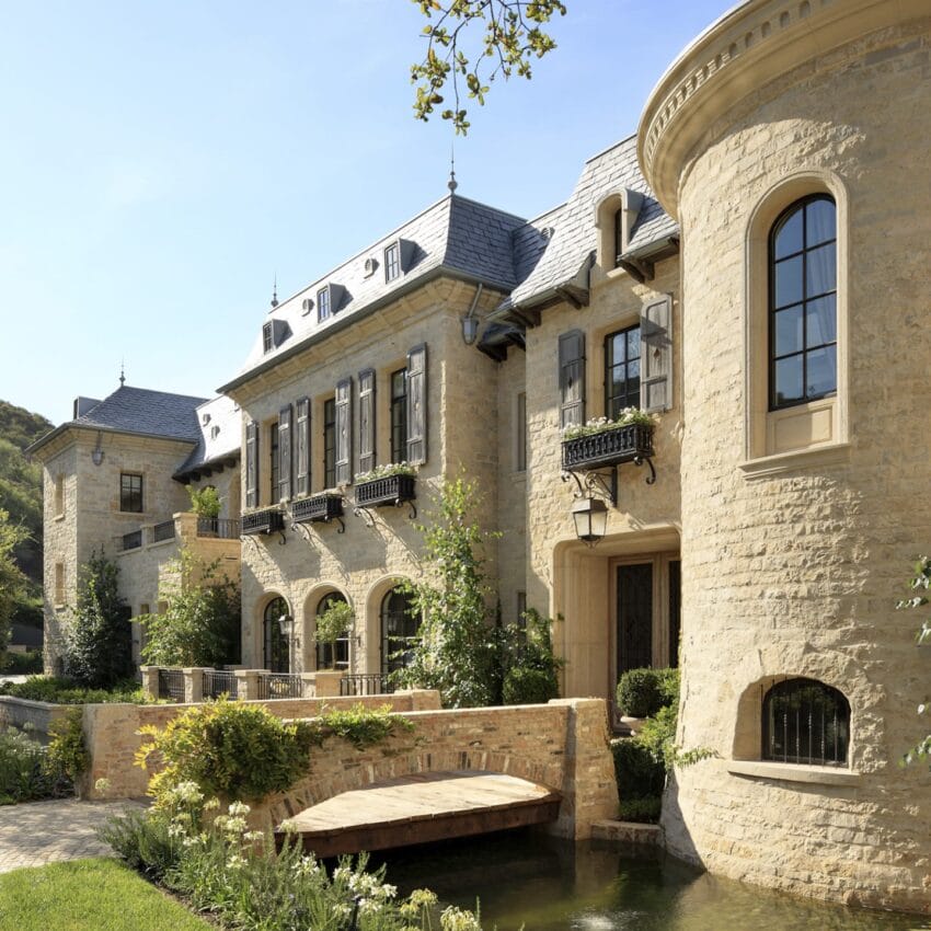 Elegant stone mansion with arched windows, turrets, and lush greenery surrounding a small wooden bridge over a pond.