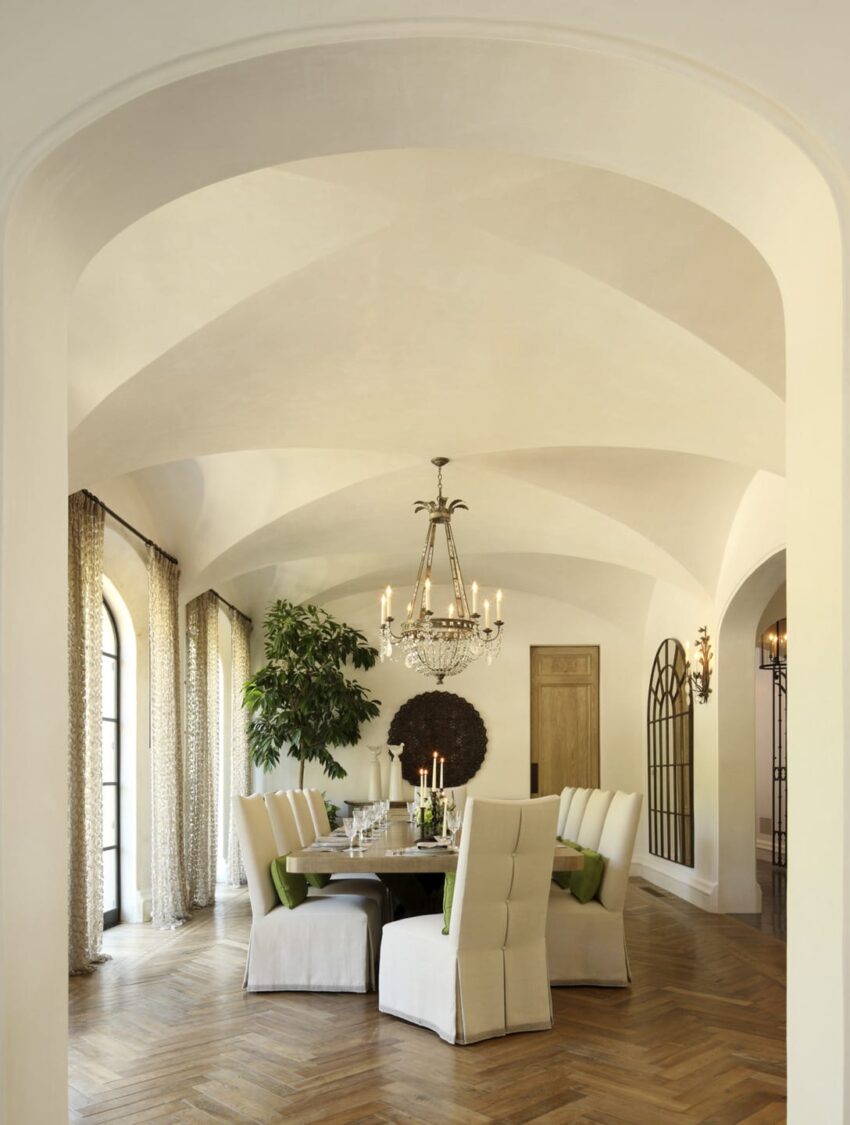 Elegant dining room with arched ceiling, chandelier, round table, white chairs, and large windows.
