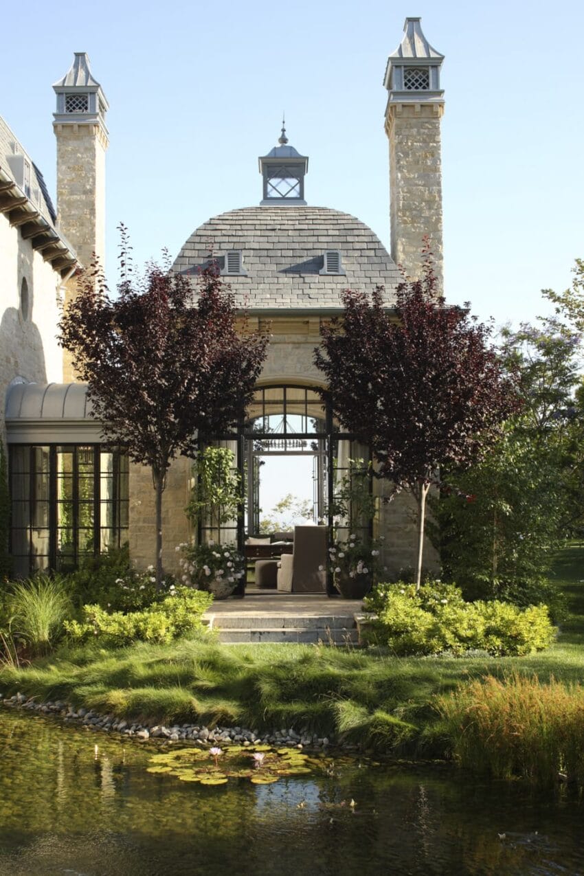 Elegant stone building with tall chimneys, surrounded by lush greenery and pond in foreground under a clear blue sky.