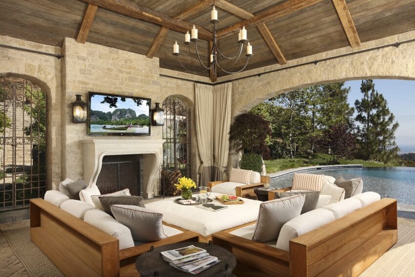 Outdoor living area with a wood-beamed ceiling, plush seating, fireplace, TV, and view of a swimming pool and garden.