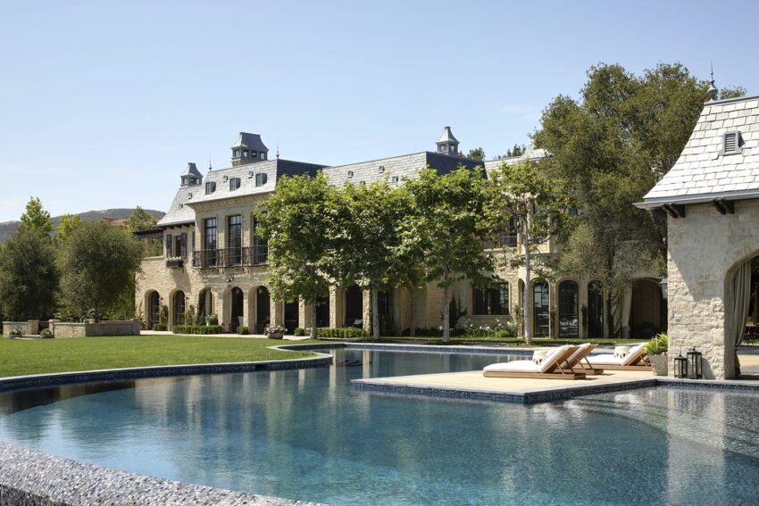 Large luxury estate with pool, surrounded by trees and manicured lawn under clear blue sky.