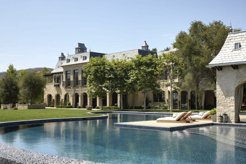 Large luxury estate with pool, surrounded by trees and manicured lawn under clear blue sky.