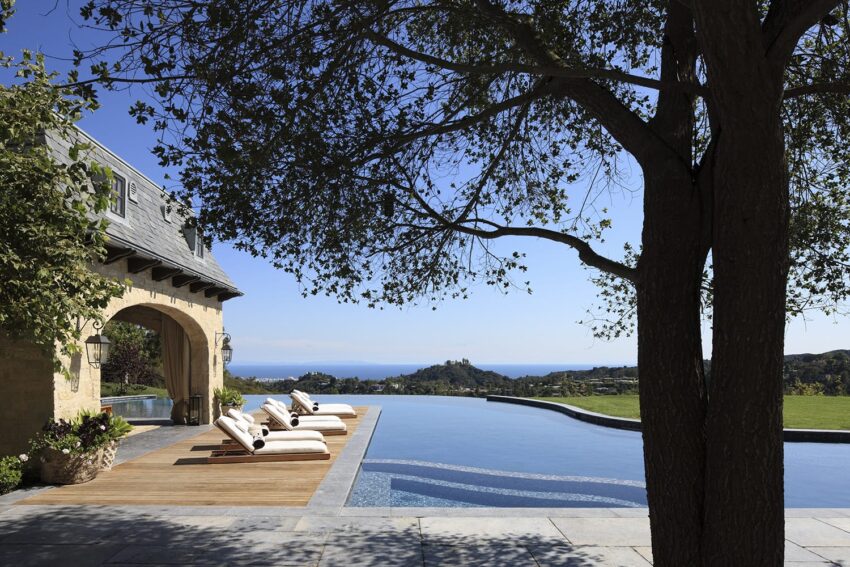 Luxury poolside with sun loungers overlooking a scenic landscape and ocean view, shaded by a large tree in the foreground.