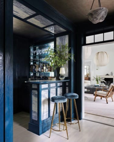 Home bar with blue and marble design, two stools, and a lamp, opening to a living room with natural light and decor.