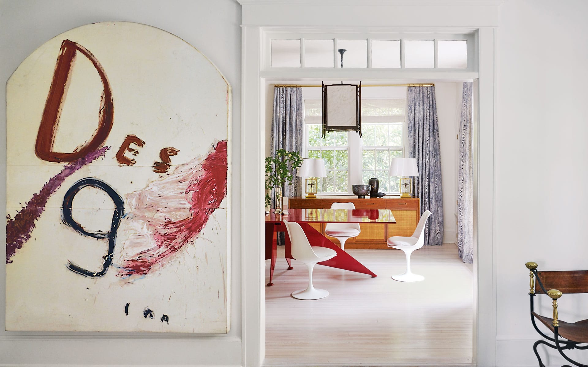 Abstract painting near a modern dining area with red table, white chairs, and floor-to-ceiling windows with blue curtains.