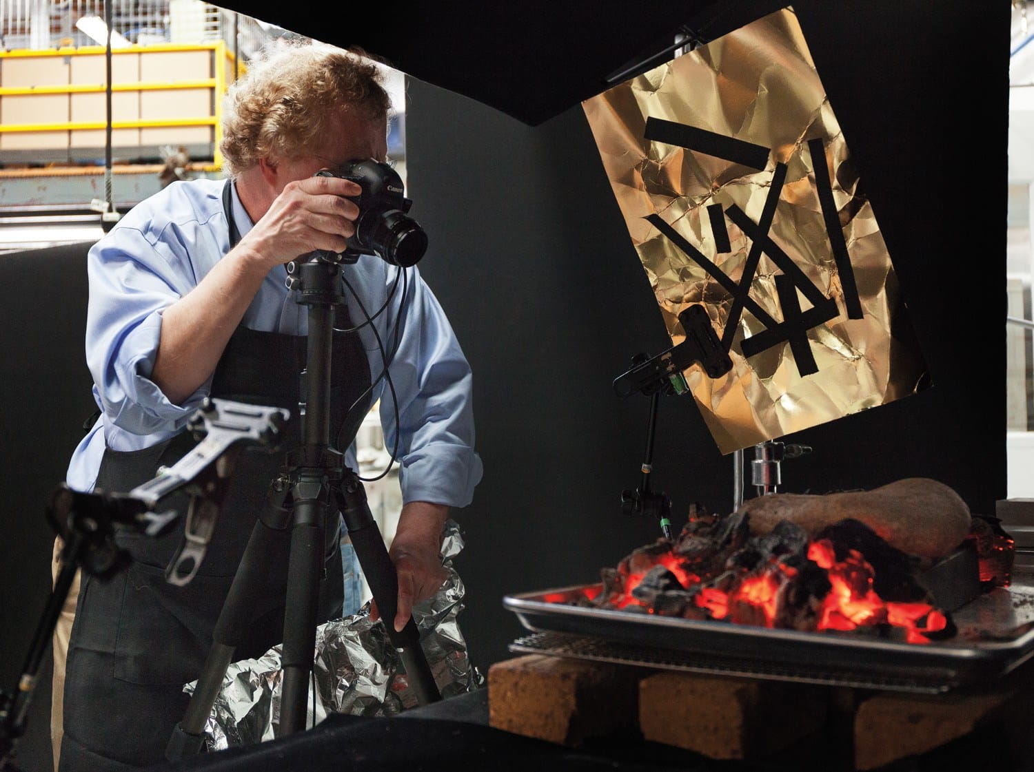 Photographer capturing an image of sizzling steak on a grill with a camera setup and lighting adjustments.