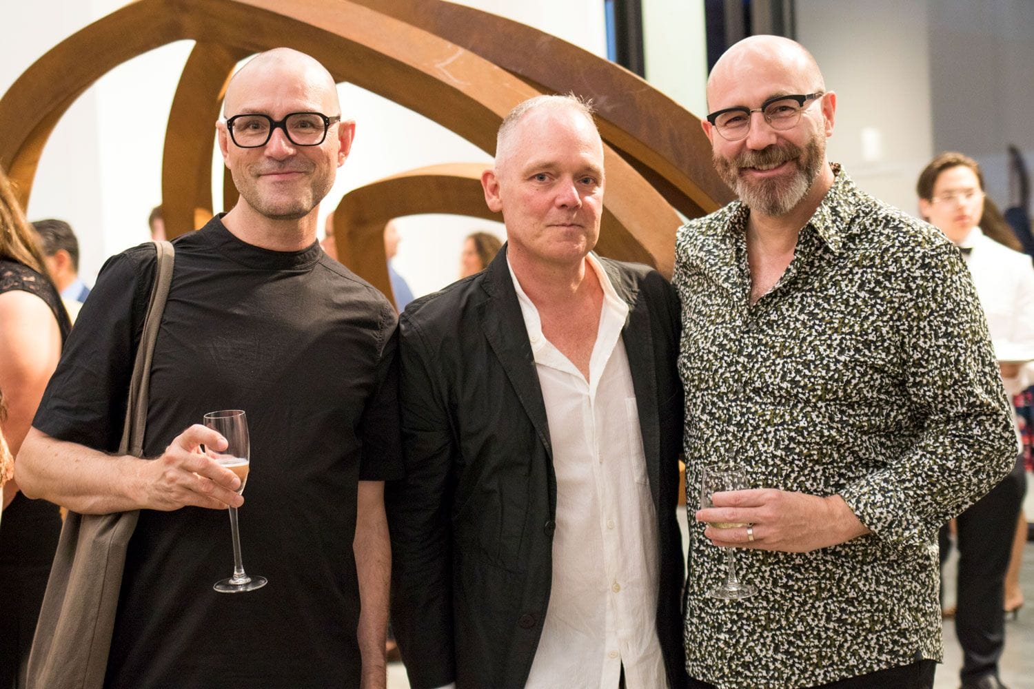 Three men standing together at an art gallery event, holding wine glasses and posing in front of a wooden sculpture.