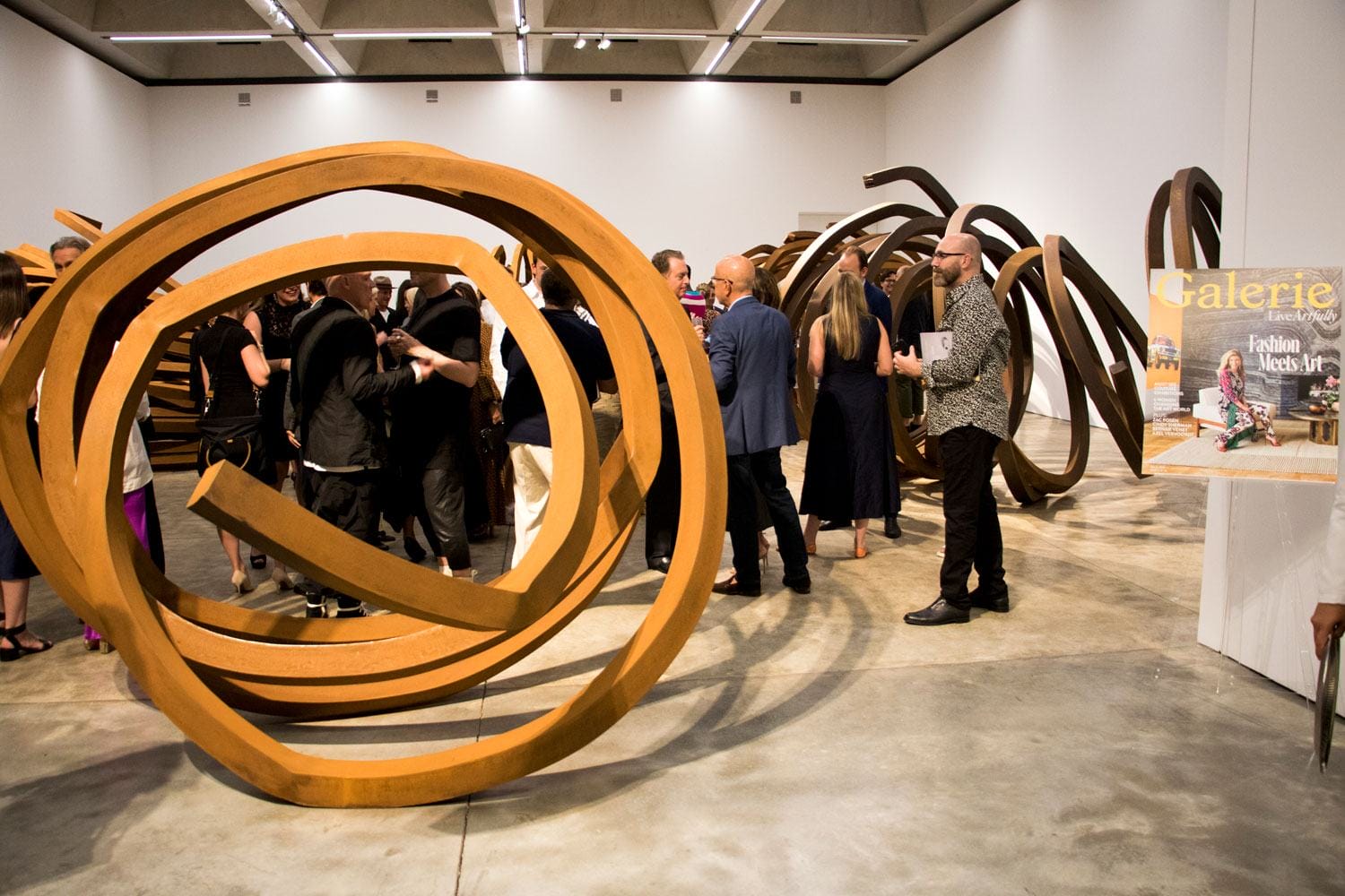 Art gallery event with people interacting around large abstract metallic sculptures displayed on a concrete floor.