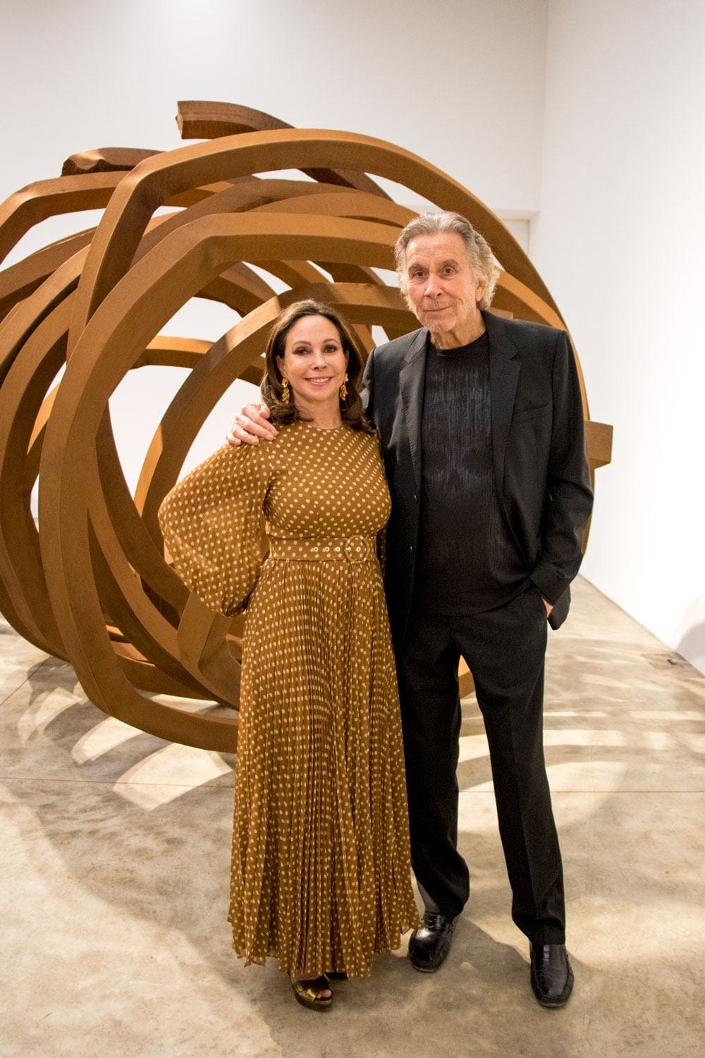 Two people standing in front of a large abstract sculpture in a gallery setting.