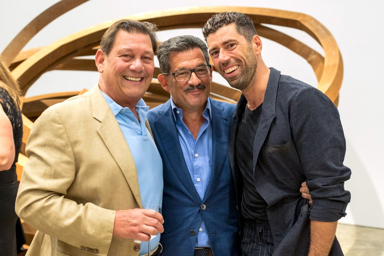 Three men smiling and posing together at an indoor event with a large wooden sculpture in the background.