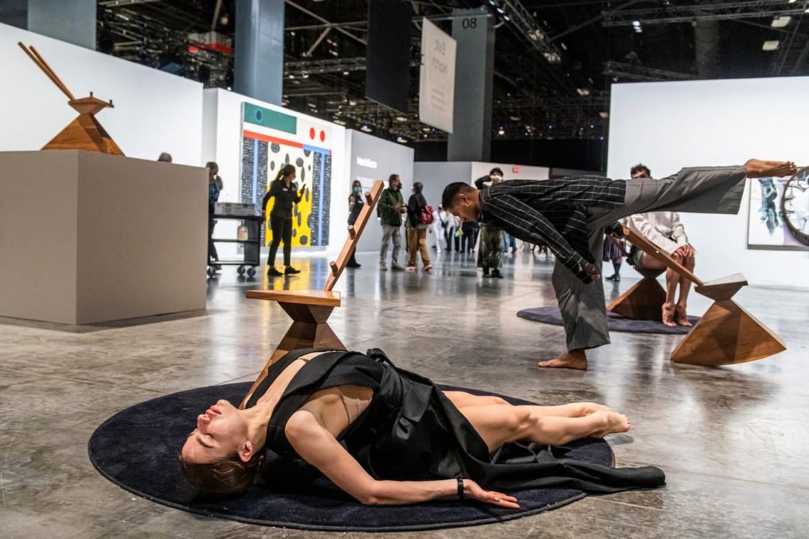 Art exhibition with performers interacting with sculptural pieces on a gallery floor, engaging an audience.