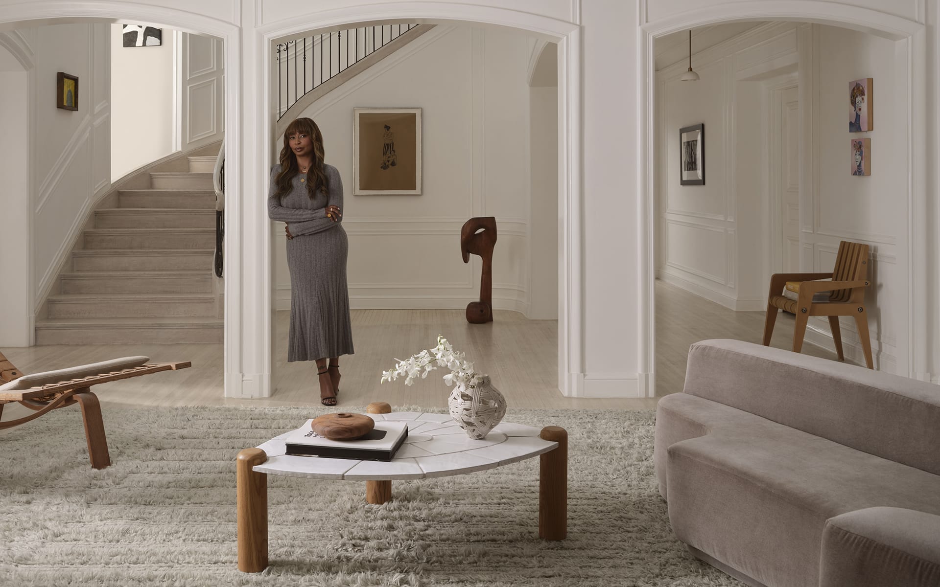 A woman stands in a modern living room with elegant decor, featuring a white rug, wooden furniture, and artwork on the walls.