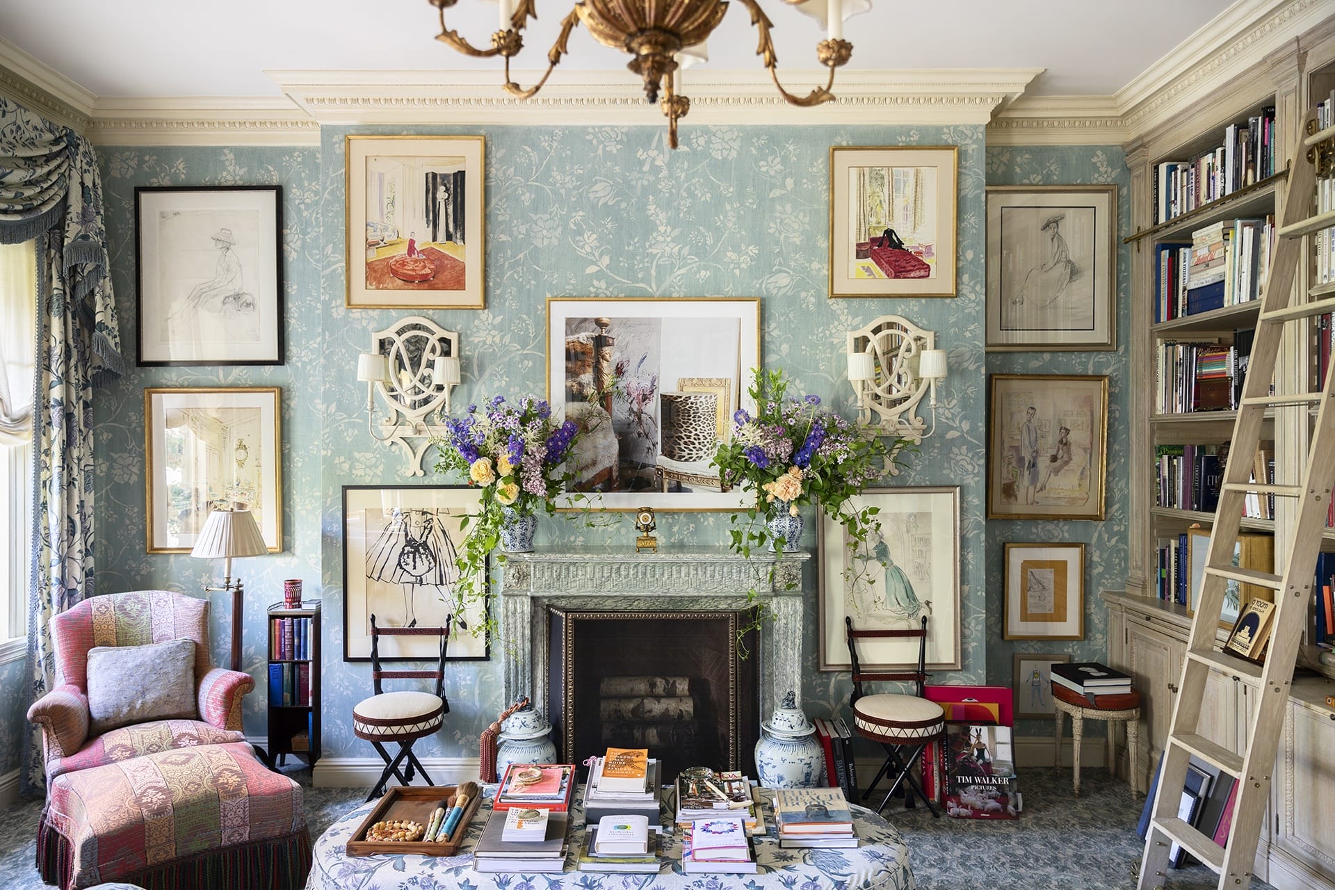 Elegant living room with floral wallpaper, art-filled walls, bookshelves, and a decorative mantelpiece adorned with flowers.