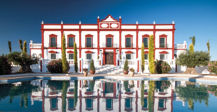 Elegant white and red mansion with tall windows, garden, and reflecting pool under a clear blue sky.