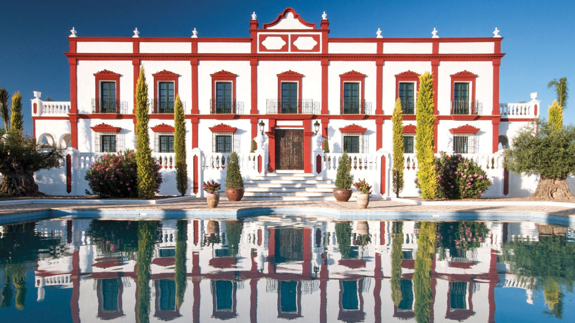 Elegant manor house with red and white facade reflected in a calm pool, surrounded by lush greenery under a clear blue sky.
