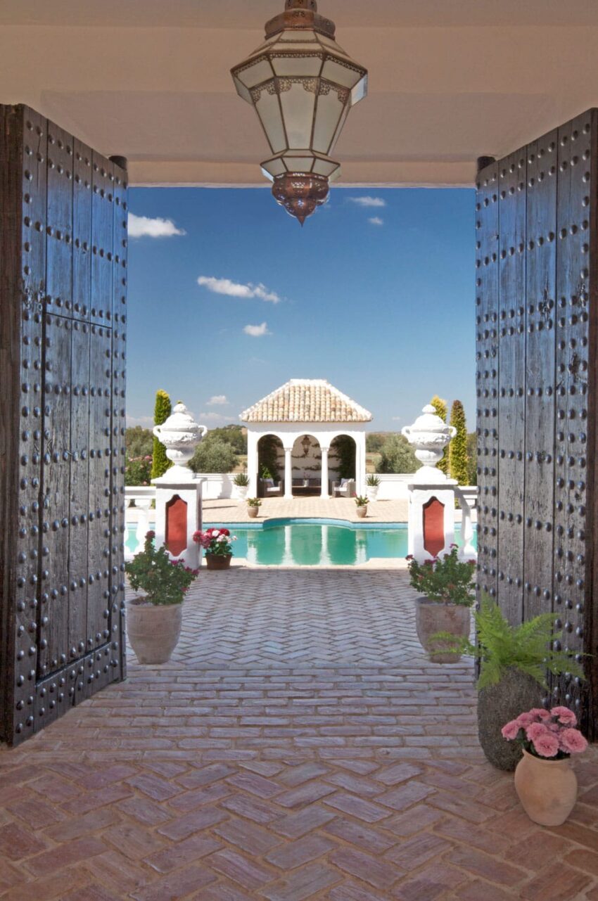 View through ornate doors to a luxurious poolside area with a small pavilion under a clear blue sky.