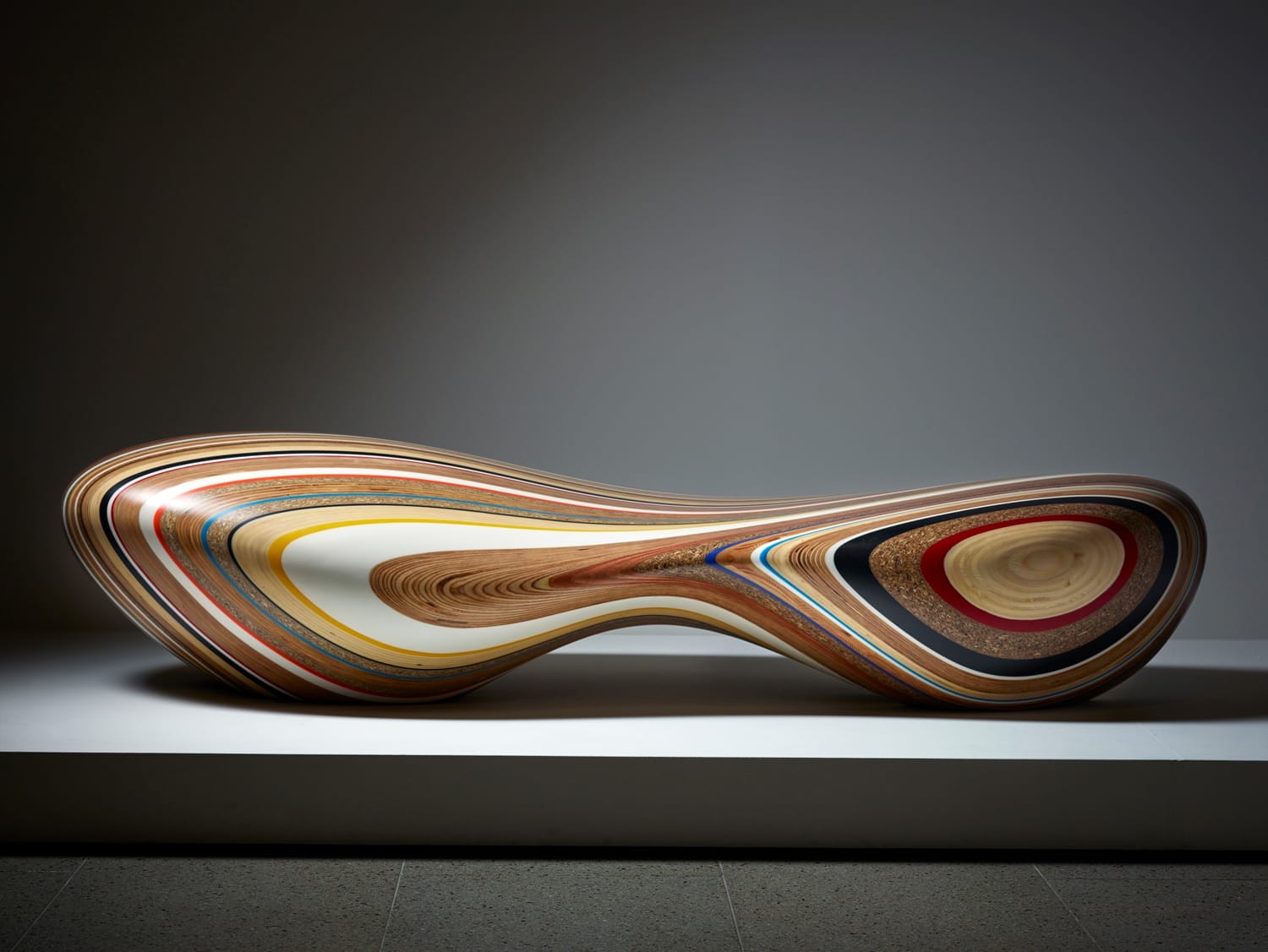 Abstract wooden sculpture with multi-colored layers, resembling a twisted hourglass, displayed on a minimalist pedestal.
