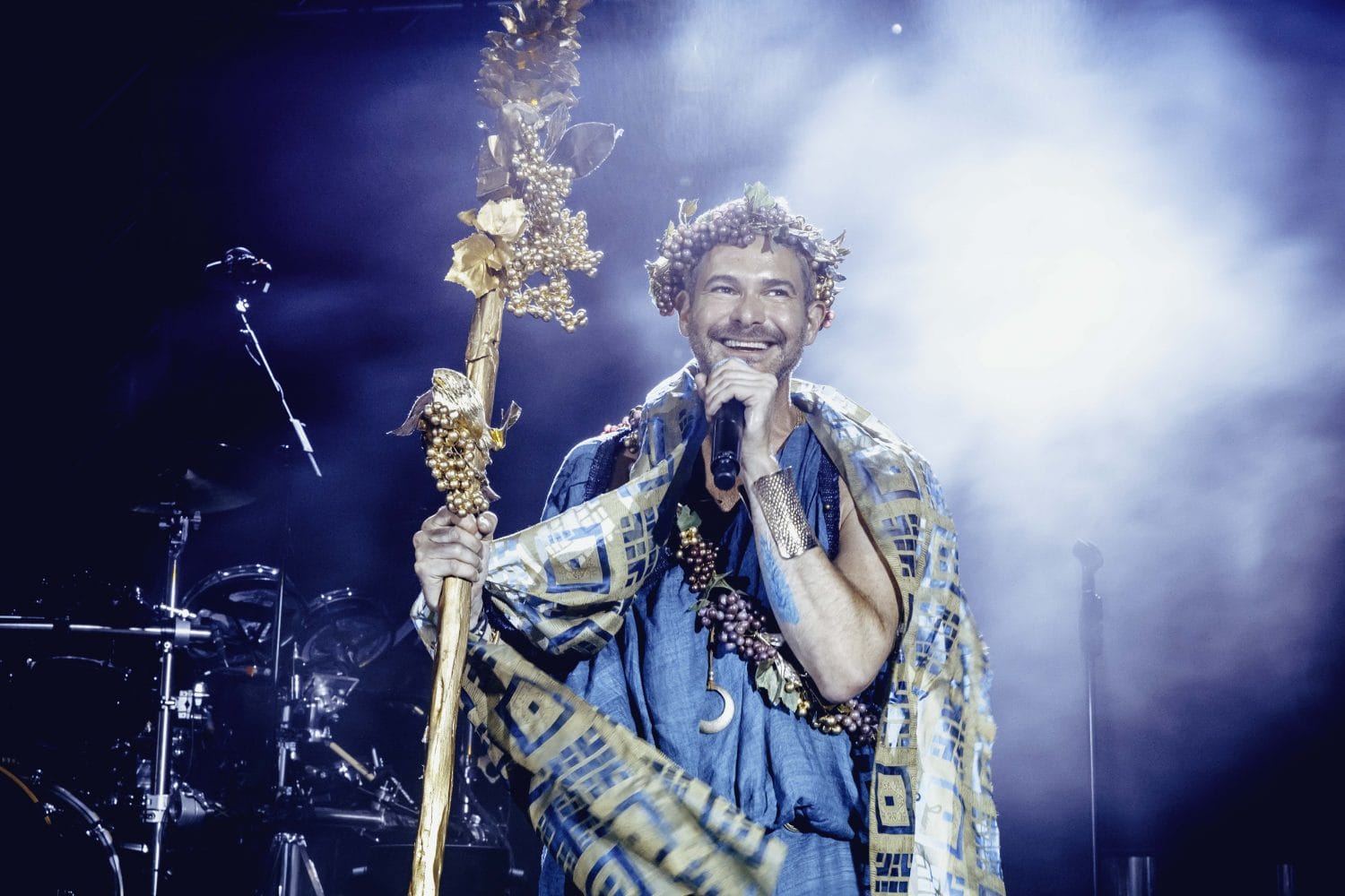 Performer in costume holding a decorated staff, standing on stage with smoke and lights in the background.