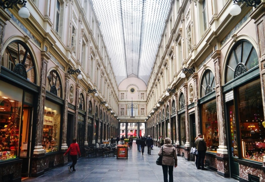 People walking inside a grand, ornate shopping arcade with a glass roof and elegant architectural details.