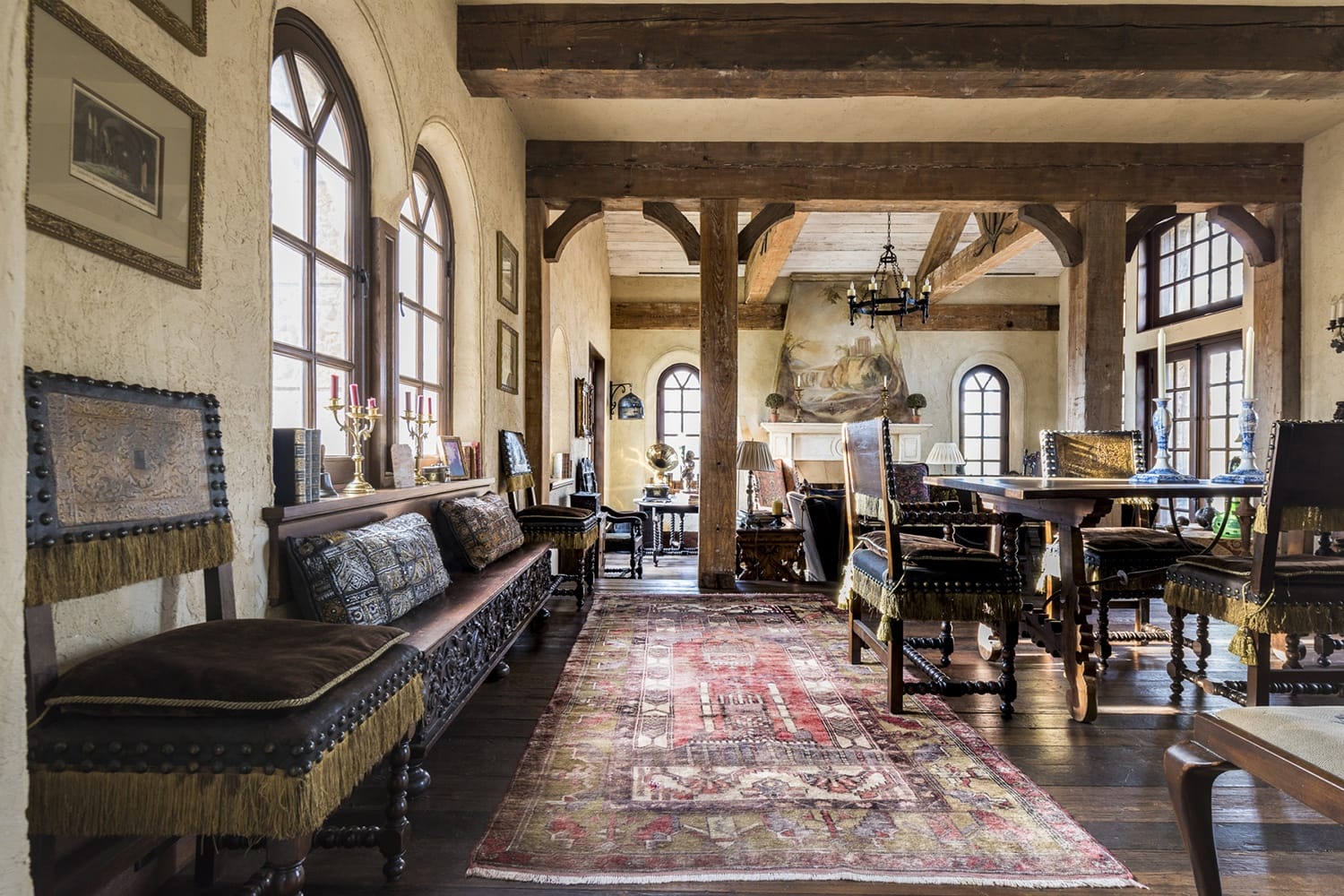 Rustic dining room with arched windows, wooden beams, vintage furniture, and a patterned rug on a wooden floor.