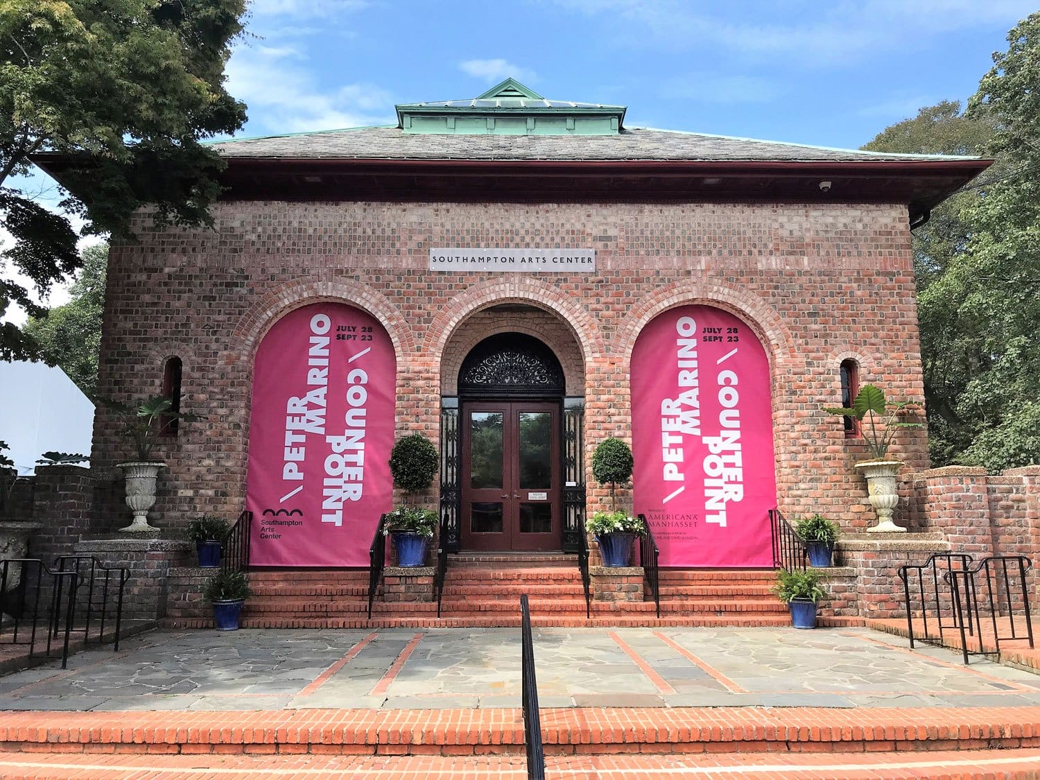 Brick building entrance with pink banners and potted plants at Southampton Arts Center under a blue sky.