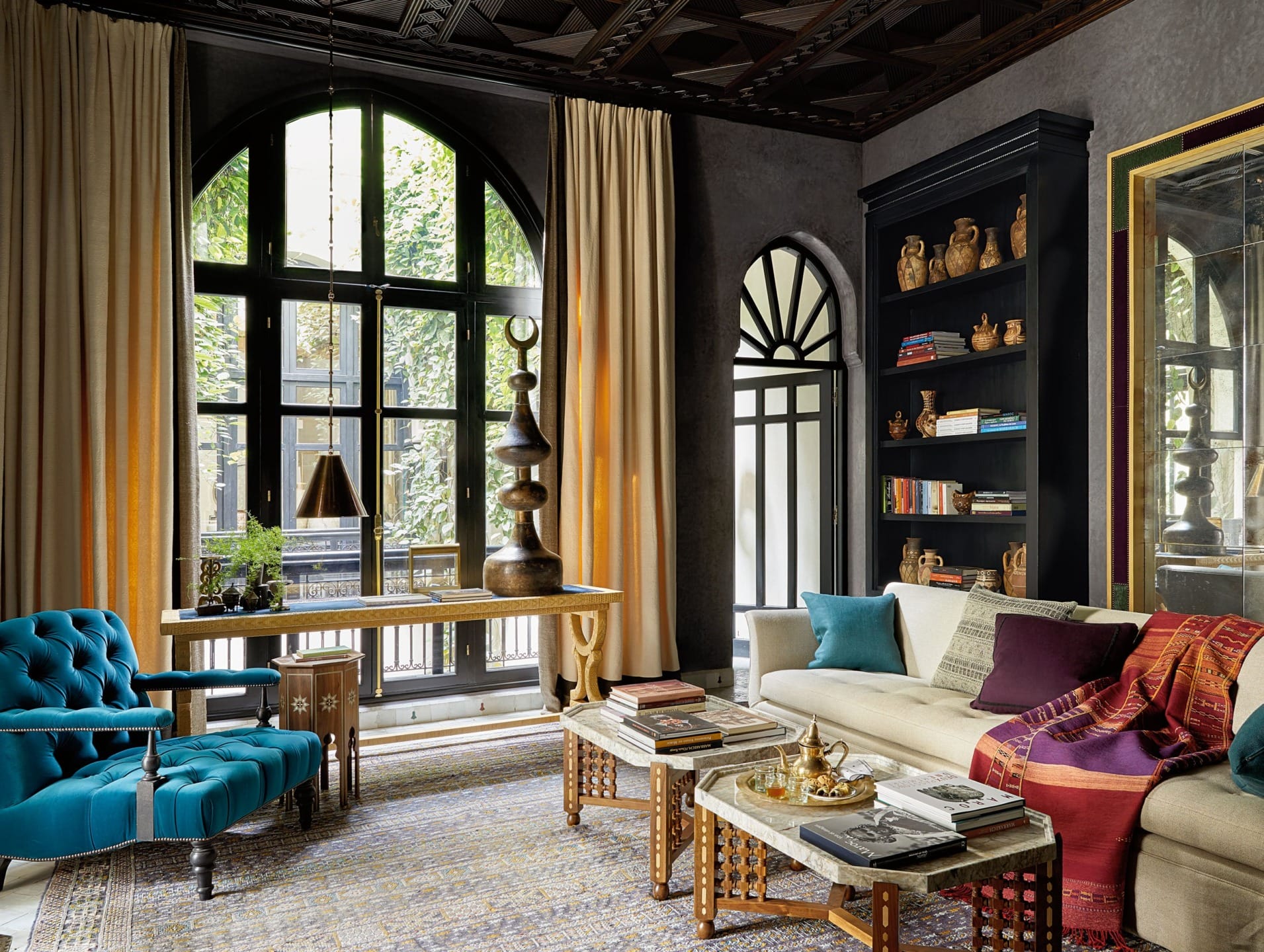 Elegant living room with arched windows, velvet armchair, sofa, decorative cushions, and bookshelves displaying pottery.