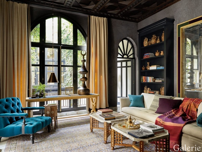 Eclectic living room with arched windows, teal chair, beige sofa, decorative shelves, and layered rugs.