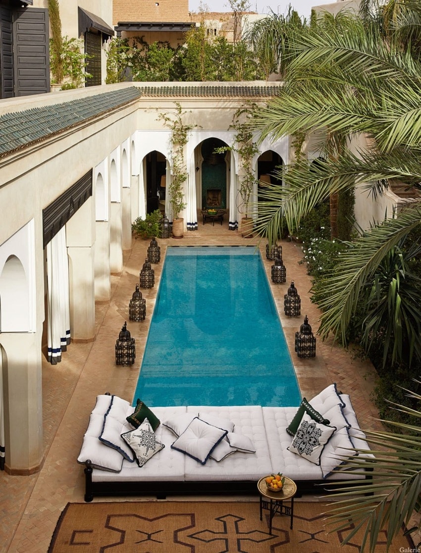 Outdoor courtyard with a swimming pool, surrounded by lanterns, palm trees, and a plush seating area with cushions.