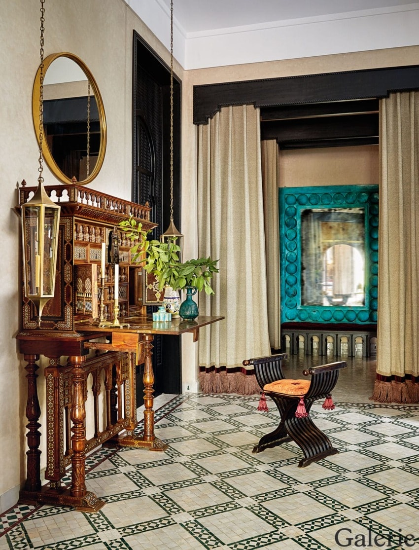 Eclectic interior with ornate wooden table, decorative floor tiles, large mirror, and unique seating in a stylish room.