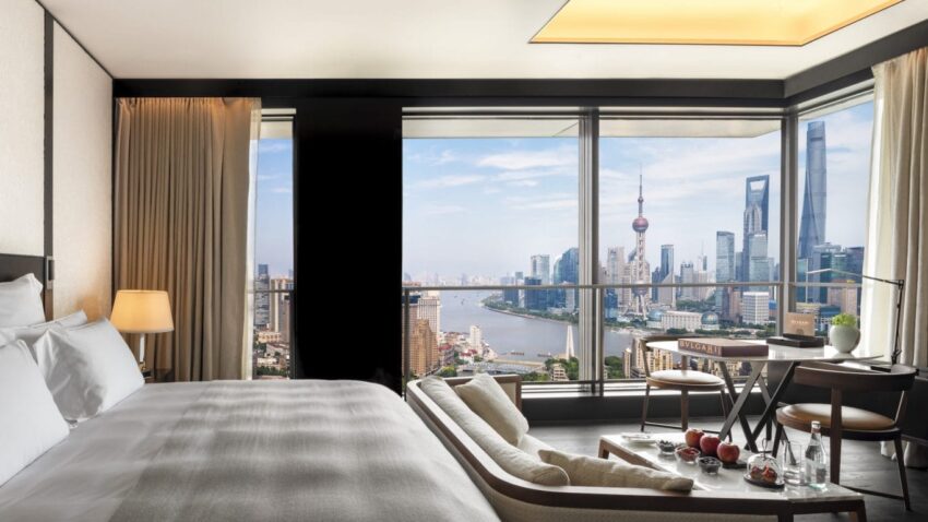 Luxury hotel room with city skyline view, large bed, floor-to-ceiling windows, and elegant furnishings.