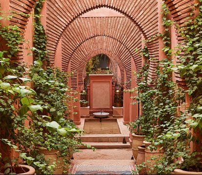 Arched pathway with red brick and green ivy leads to a small fountain in a serene courtyard garden setting.