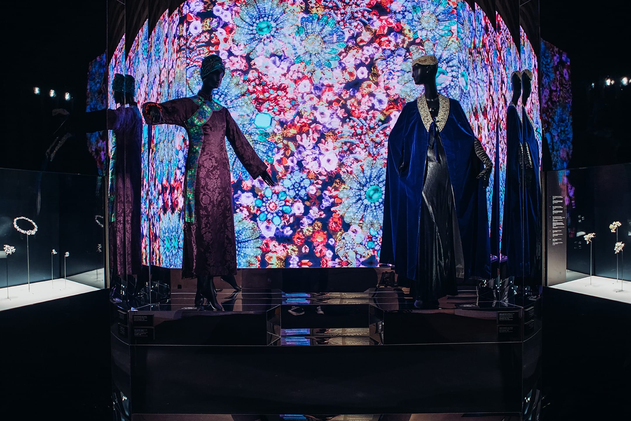Three mannequins in elegant gowns, posed in front of a vibrant, colorful digital floral display at an exhibition.