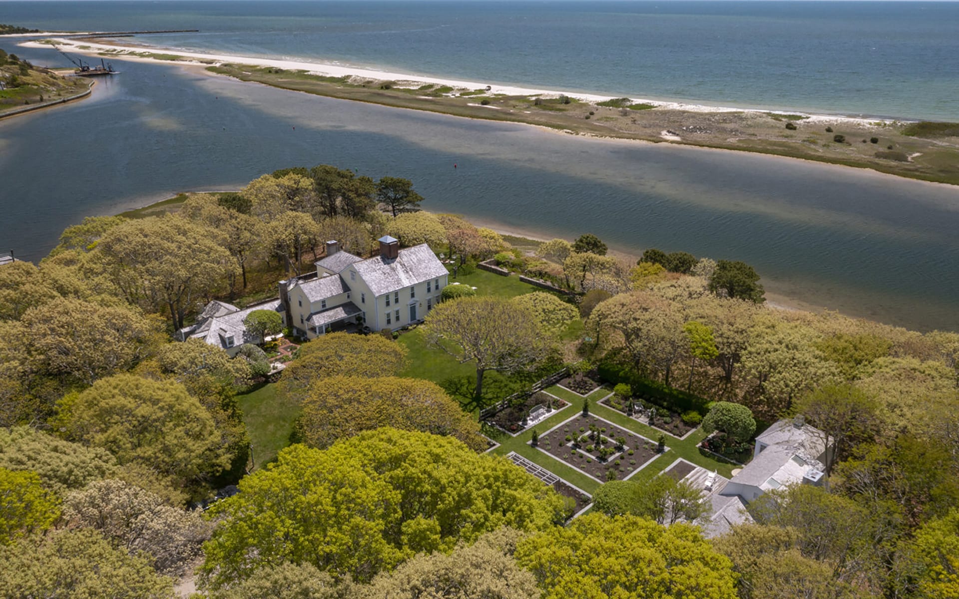 Aerial view of a coastal mansion surrounded by lush gardens and trees, near a river and ocean.
