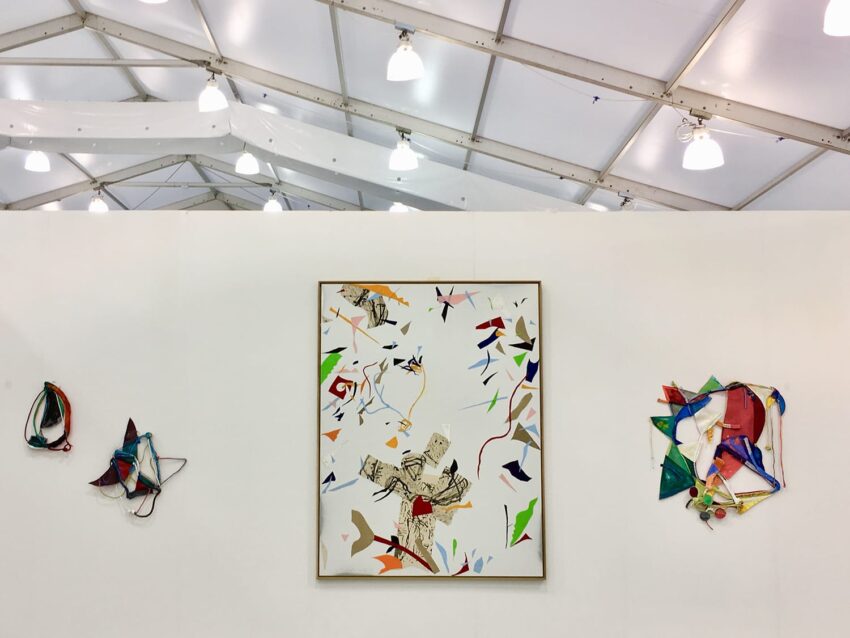 Art gallery display featuring abstract paintings and sculptures on a white wall under a bright, modern tent ceiling.