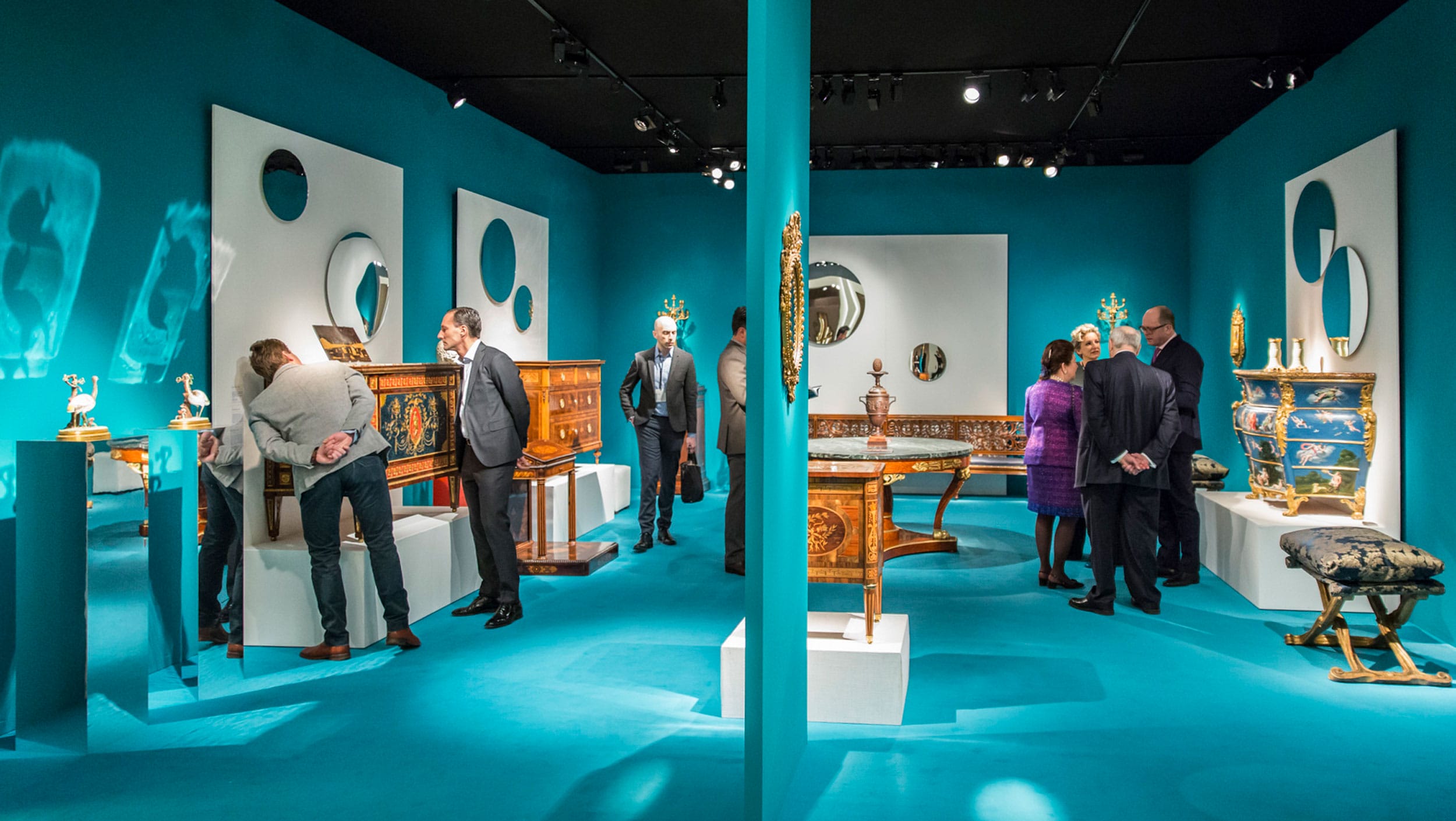 People viewing antique furniture in a brightly lit exhibition room with turquoise walls and mirrors.