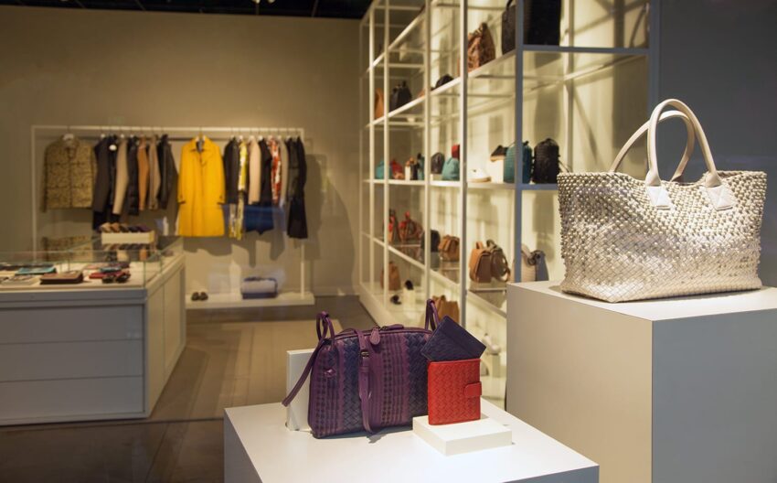 Fashion store interior with stylish handbags on display in the foreground and clothes hanging on racks in the background.
