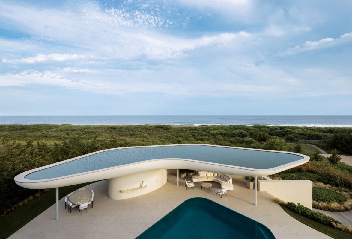 Modern house with a unique curved roof and infinity pool overlooking the ocean, surrounded by lush greenery under a blue sky.