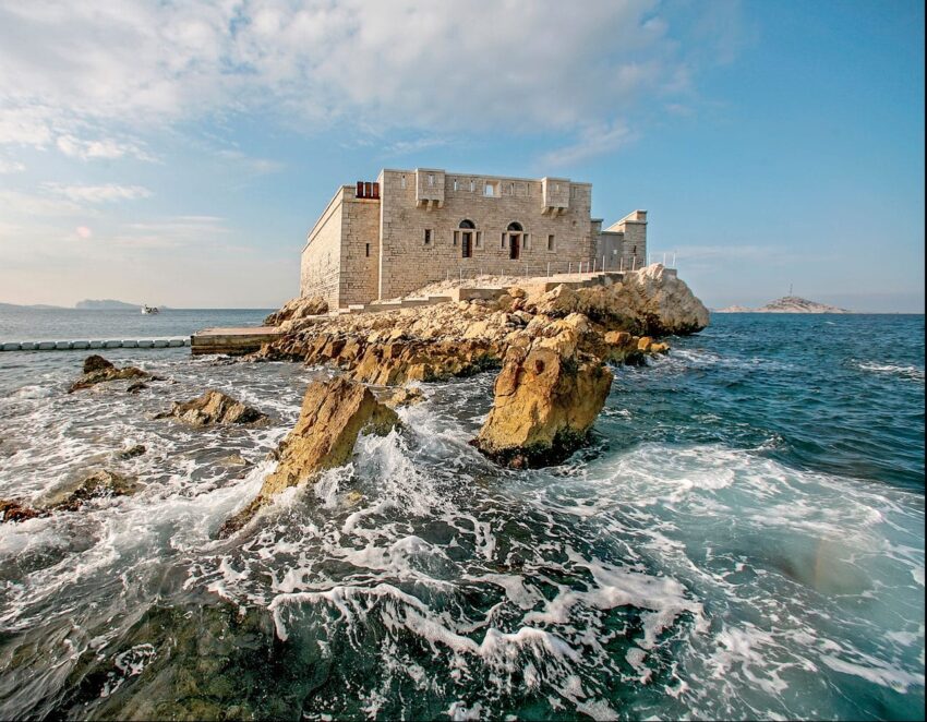 Historic stone fortress on a rocky island surrounded by ocean waves under a partly cloudy blue sky.