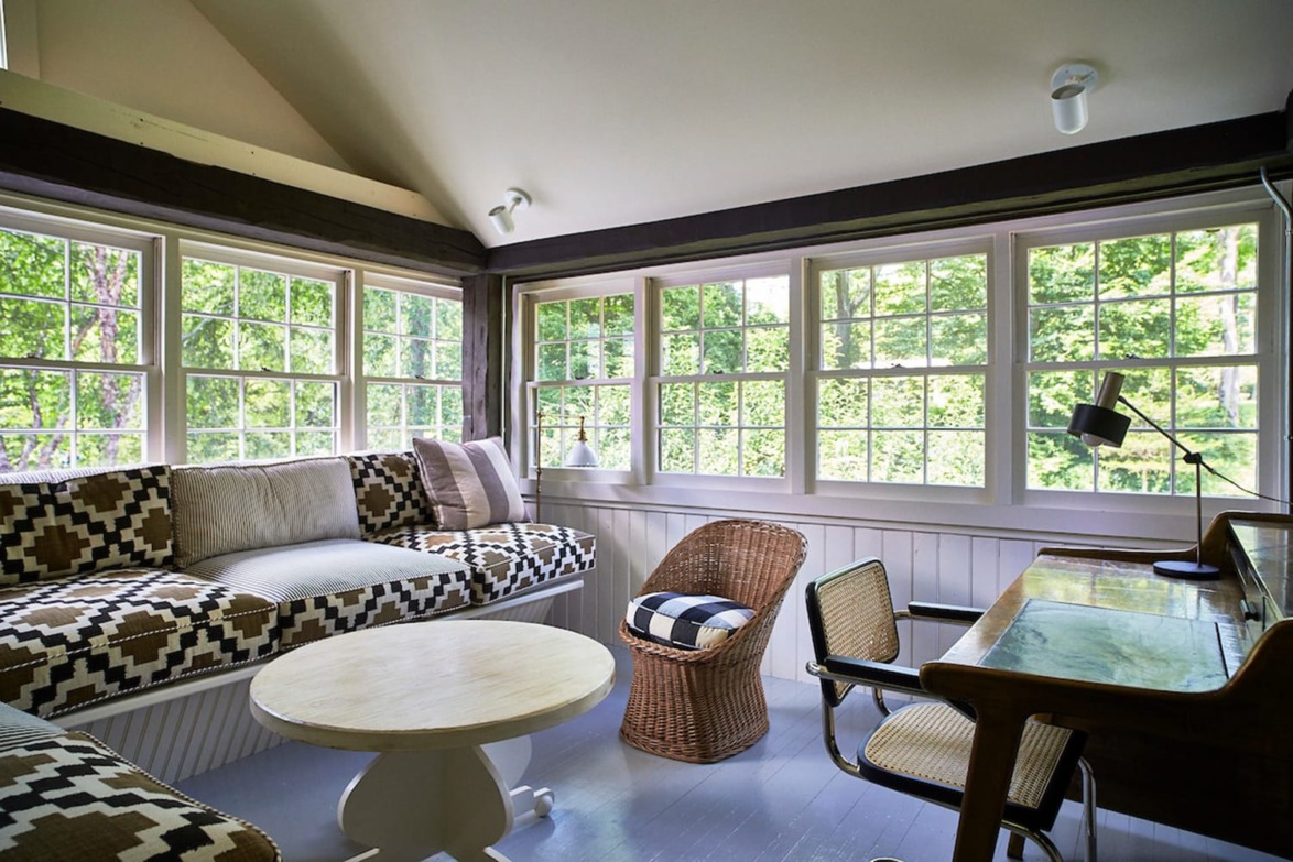 Cozy sunroom with patterned cushions, wicker chairs, and large windows overlooking lush green trees.