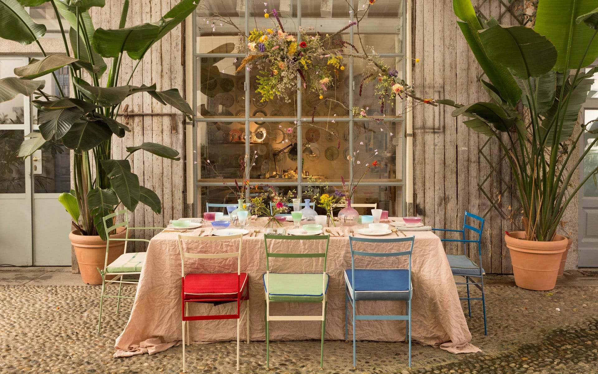 Eclectic outdoor dining setup with mismatched chairs, floral centerpiece, surrounded by large potted plants in a rustic setting.