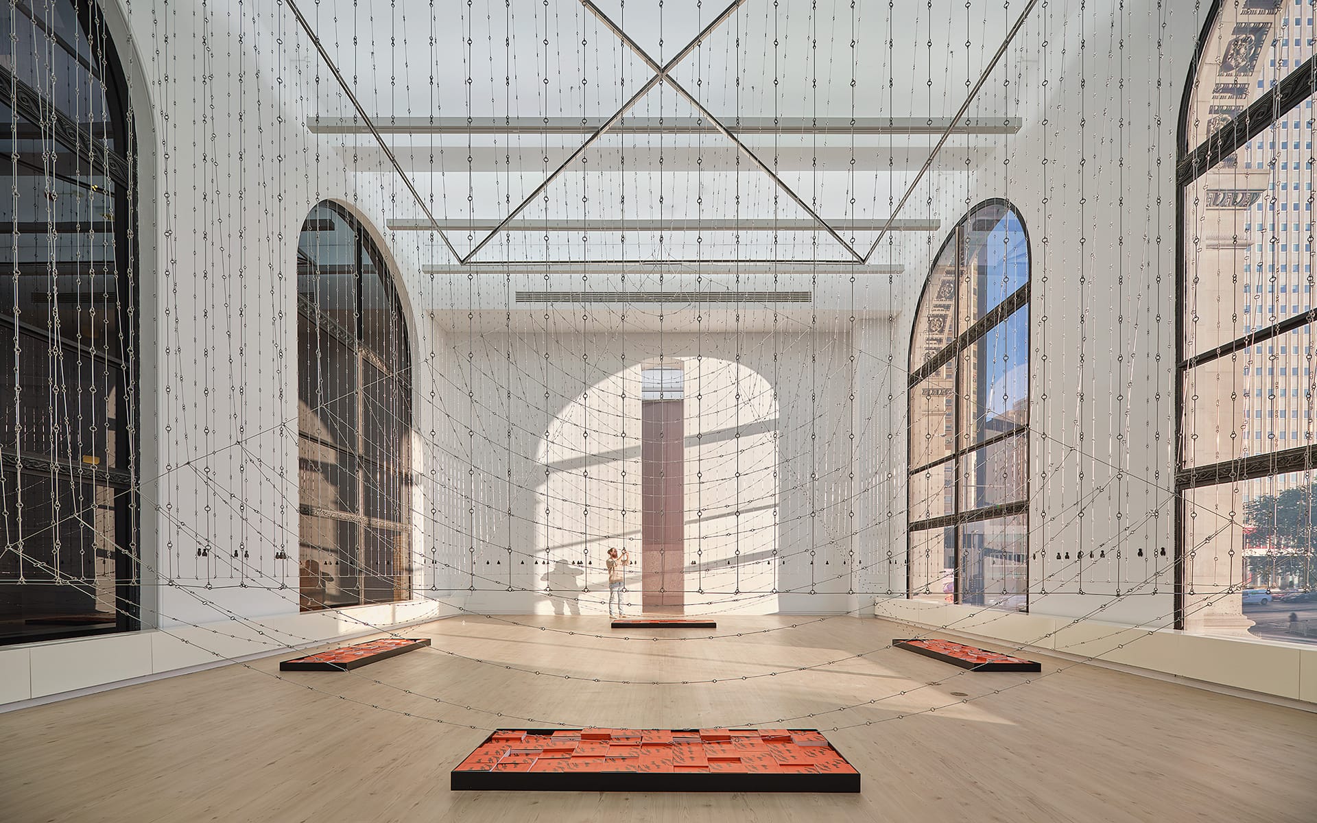 Modern art installation with hanging wires and rectangular floor pieces in a bright, spacious gallery with large arched windows.