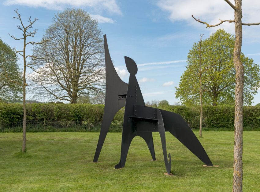 Modern abstract sculpture on grassy field surrounded by trees under a partly cloudy sky.