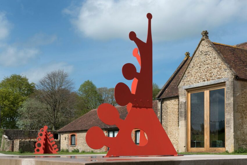 Red abstract sculptures outside rustic stone buildings under a blue sky.