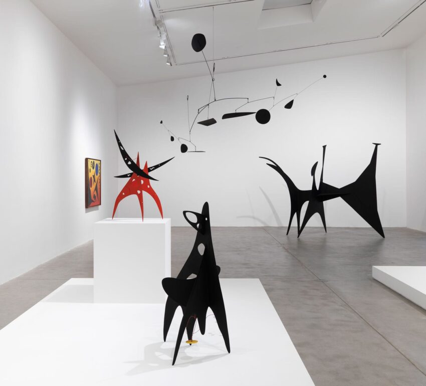 Modern art sculptures in a gallery with black and red abstract pieces and a hanging mobile on a white background.
