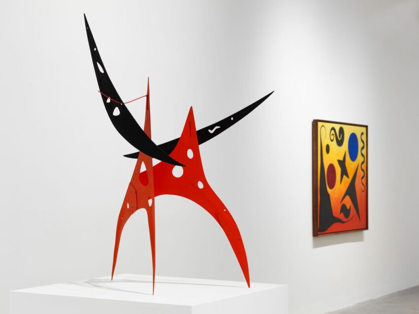 Red and black abstract sculpture with curved shapes in a gallery setting, painting on the wall in the background.