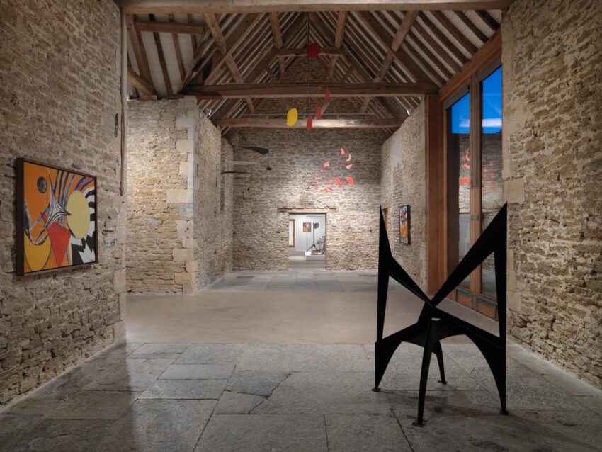 Art gallery interior with stone walls, wooden beams, abstract sculptures, colorful paintings, and large windows.