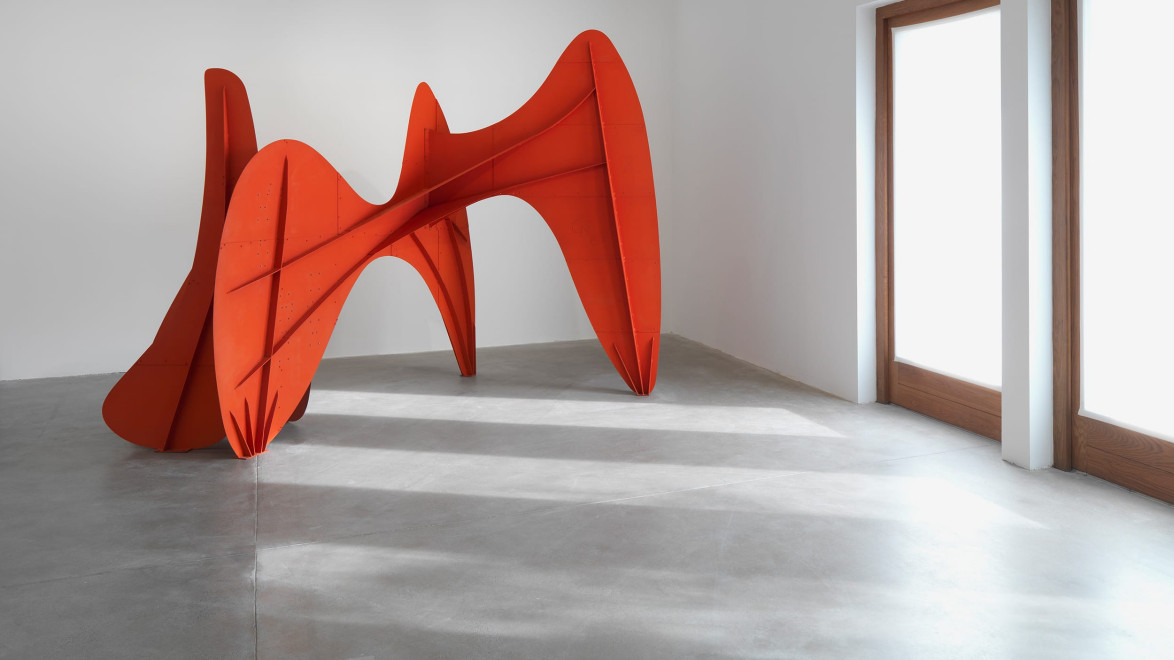 Red abstract sculpture in a modern gallery space with concrete floor and natural light from large windows.