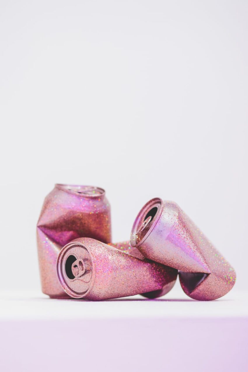 Three crushed, glittery pink soda cans placed on a white surface against a plain background.