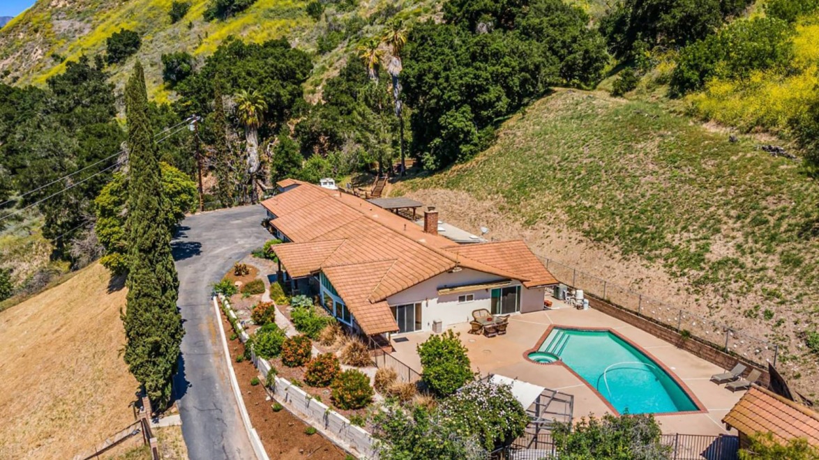 Aerial view of a house with a terracotta roof, surrounded by lush greenery and hills, featuring a swimming pool and driveway.