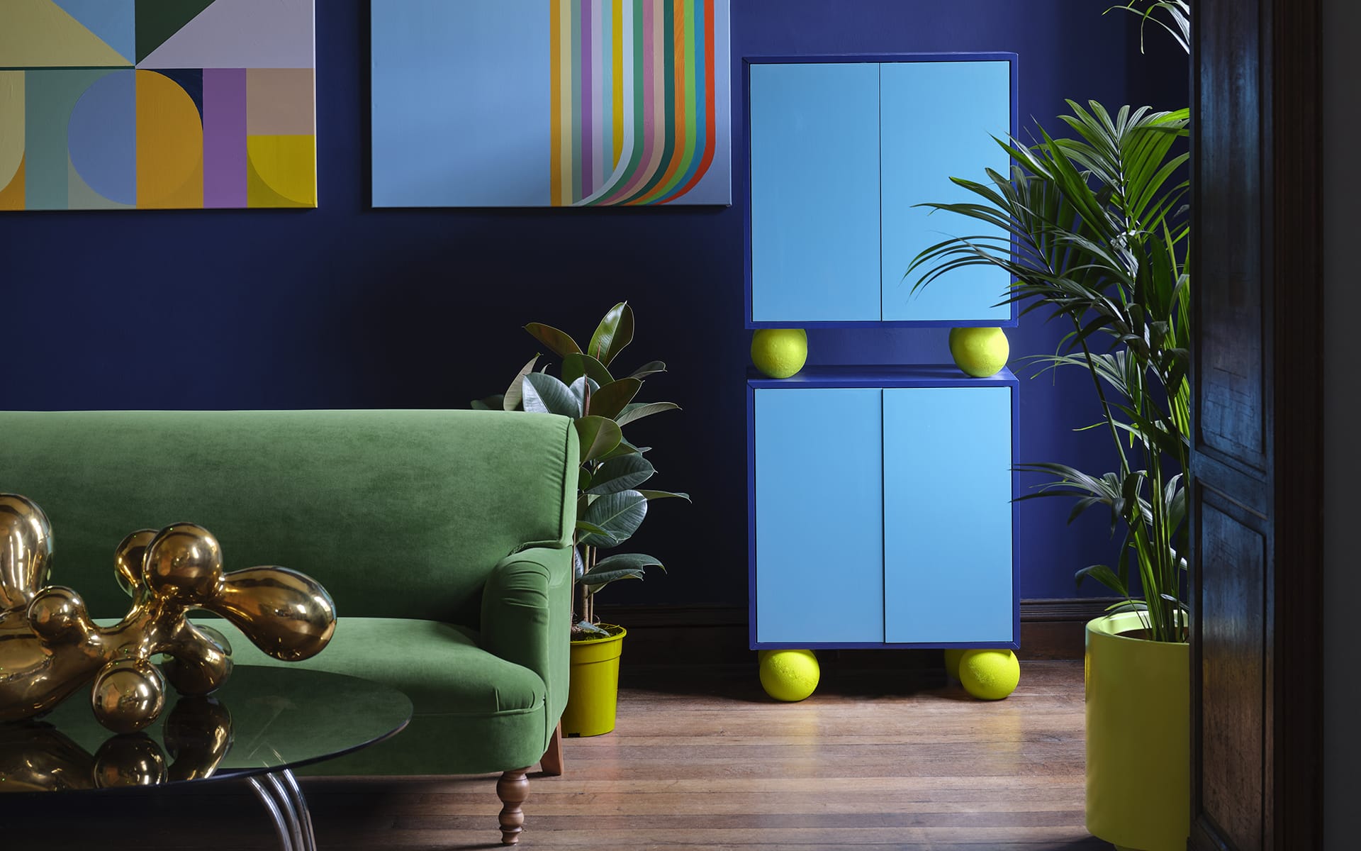 Colorful modern living room with green sofa, abstract art, blue cabinet, and large plants against a dark blue wall.